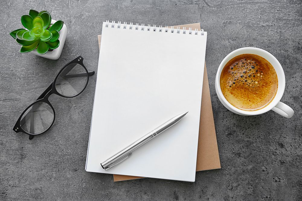 A notebook , pen , glasses , and a cup of coffee on a table.