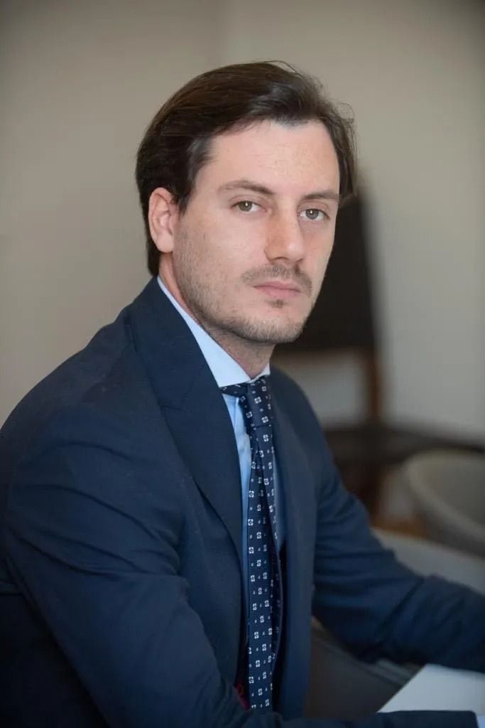 Uomo in giacca e cravatta, che guarda direttamente la telecamera, in un interno.