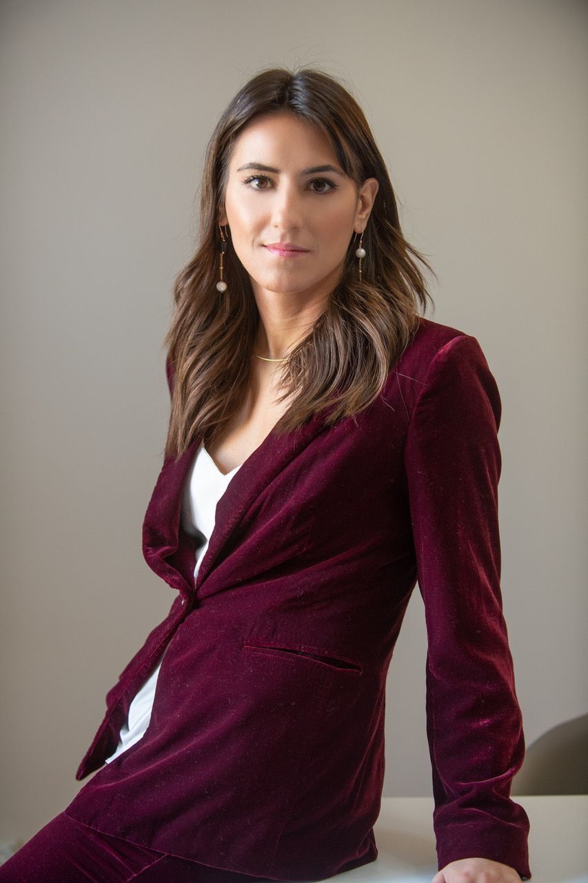 Una donna con una giacca bordeaux è appoggiata a un tavolo e guarda la telecamera. Sfondo neutro.