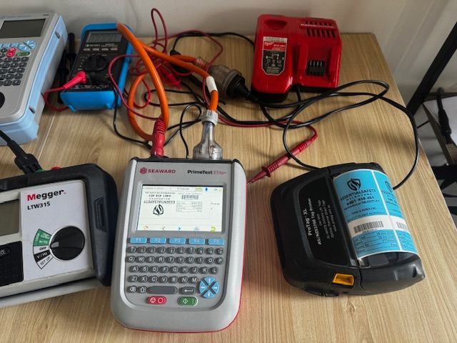 A person using a clamp meter to test wires in an electrical panel. Electrical testing equipment connected to a battery pack on a wooden table. Includes a multi-meter and charger.