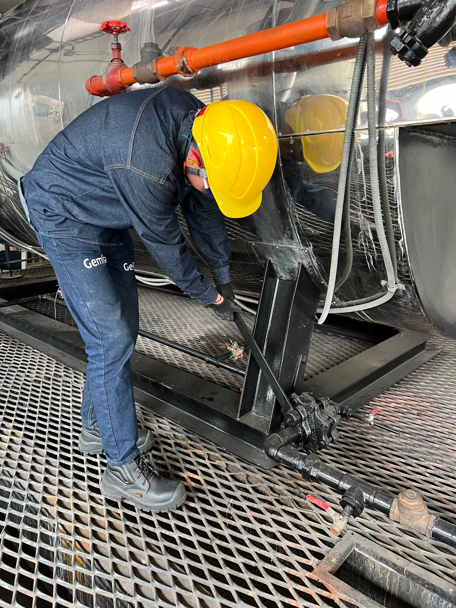 Trabajador con casco amarillo y mono azul repara equipo industrial cerca de una rejilla de metal.