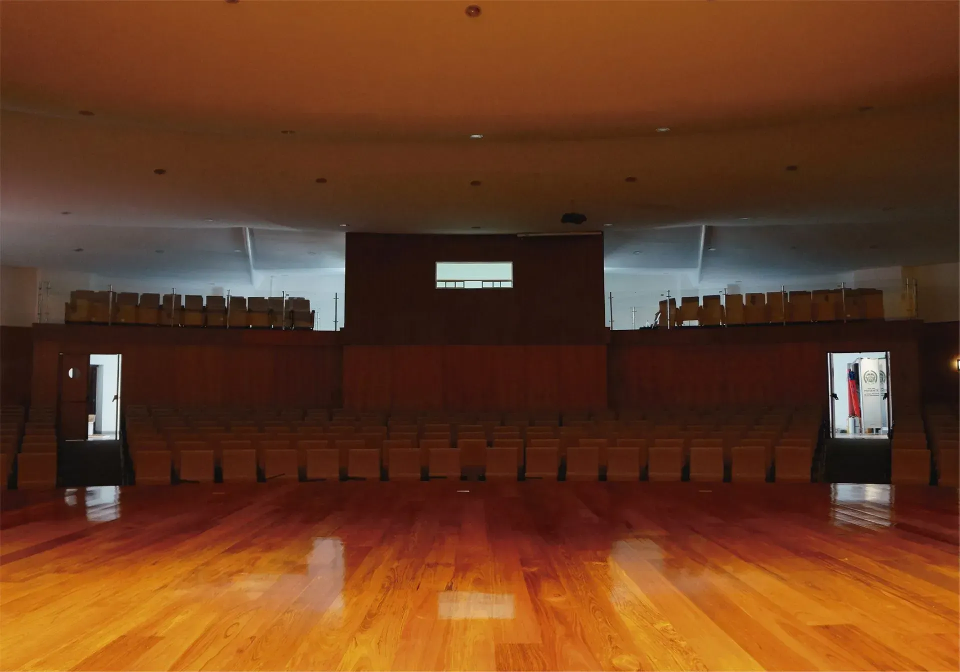Auditorio vacío con suelo de madera y escenario. Filas de asientos, puertas y una paleta de colores neutros.