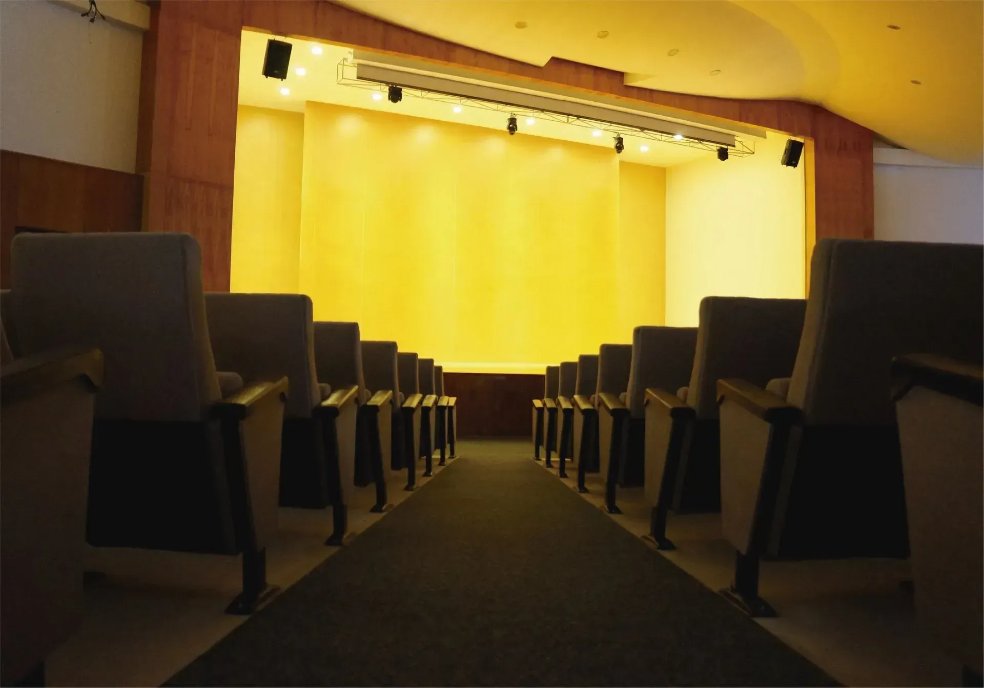 Auditorio vacío con filas de asientos frente a un escenario amarillo brillantemente iluminado, visto desde la última fila.