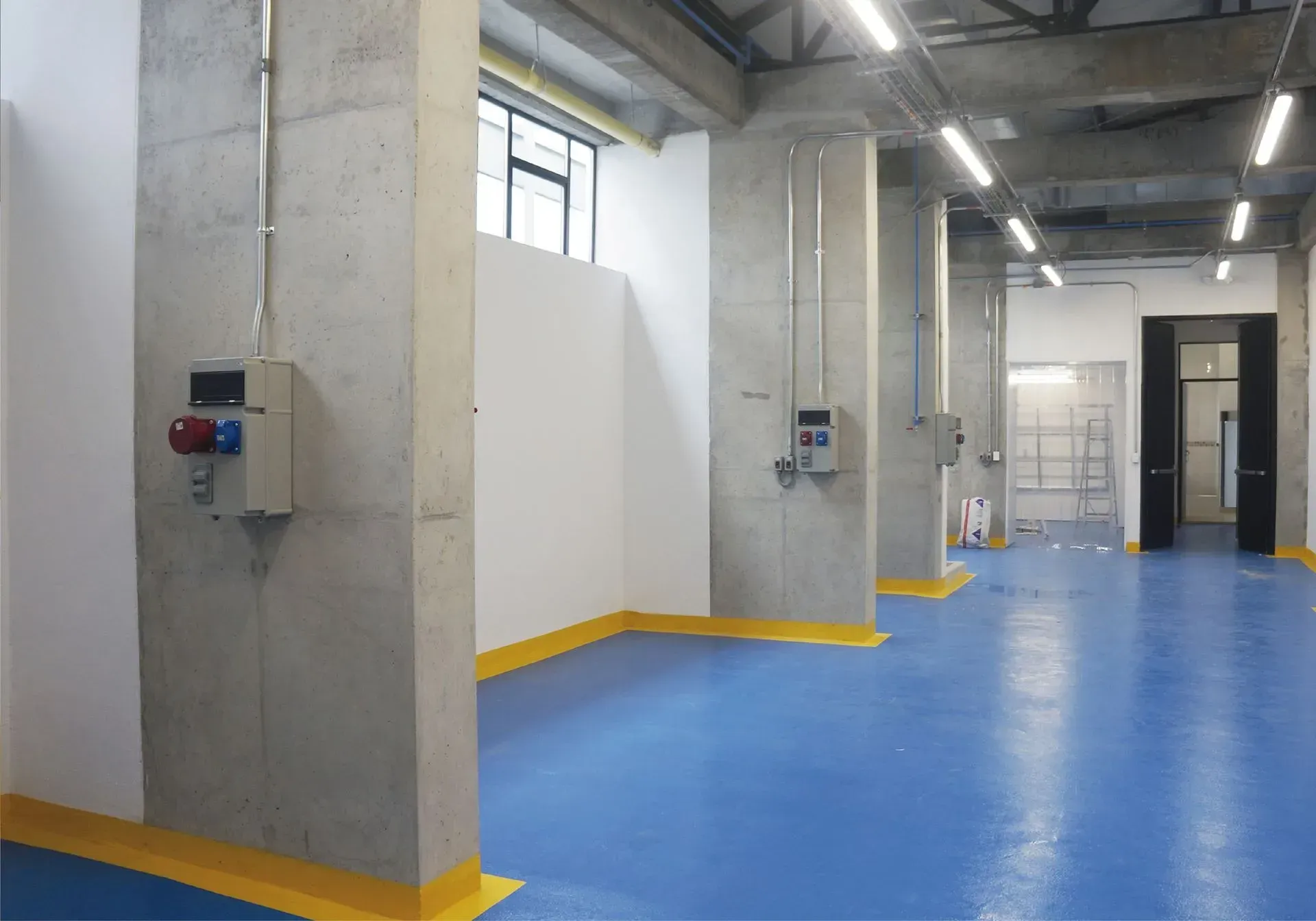 Una habitación moderna y abierta con suelo azul, columnas de hormigón y cajas eléctricas.