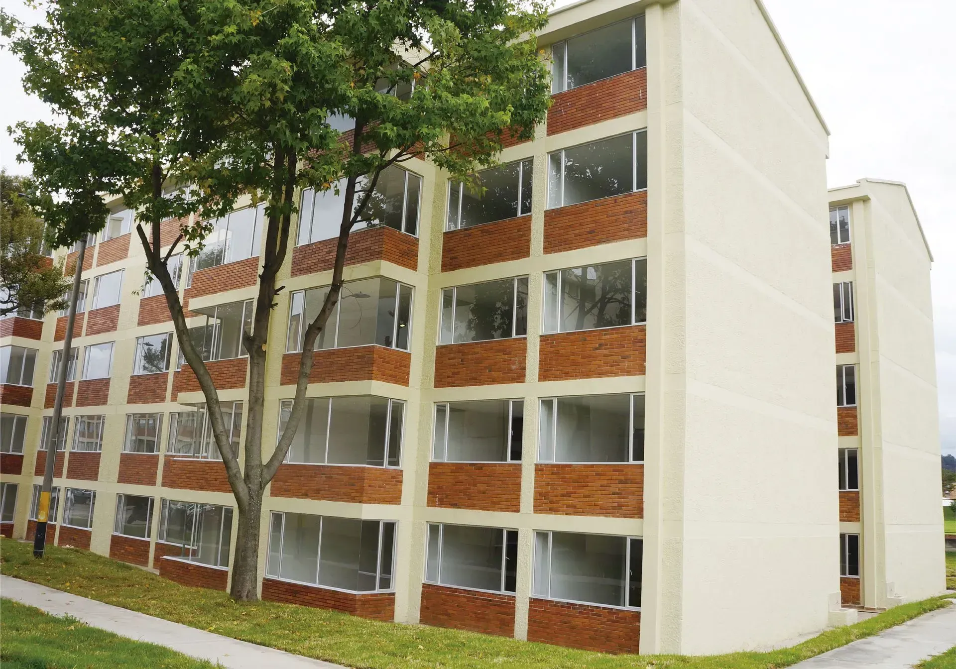 Edificio de apartamentos de varios pisos de ladrillo y beige con grandes ventanales y un árbol alto al frente.