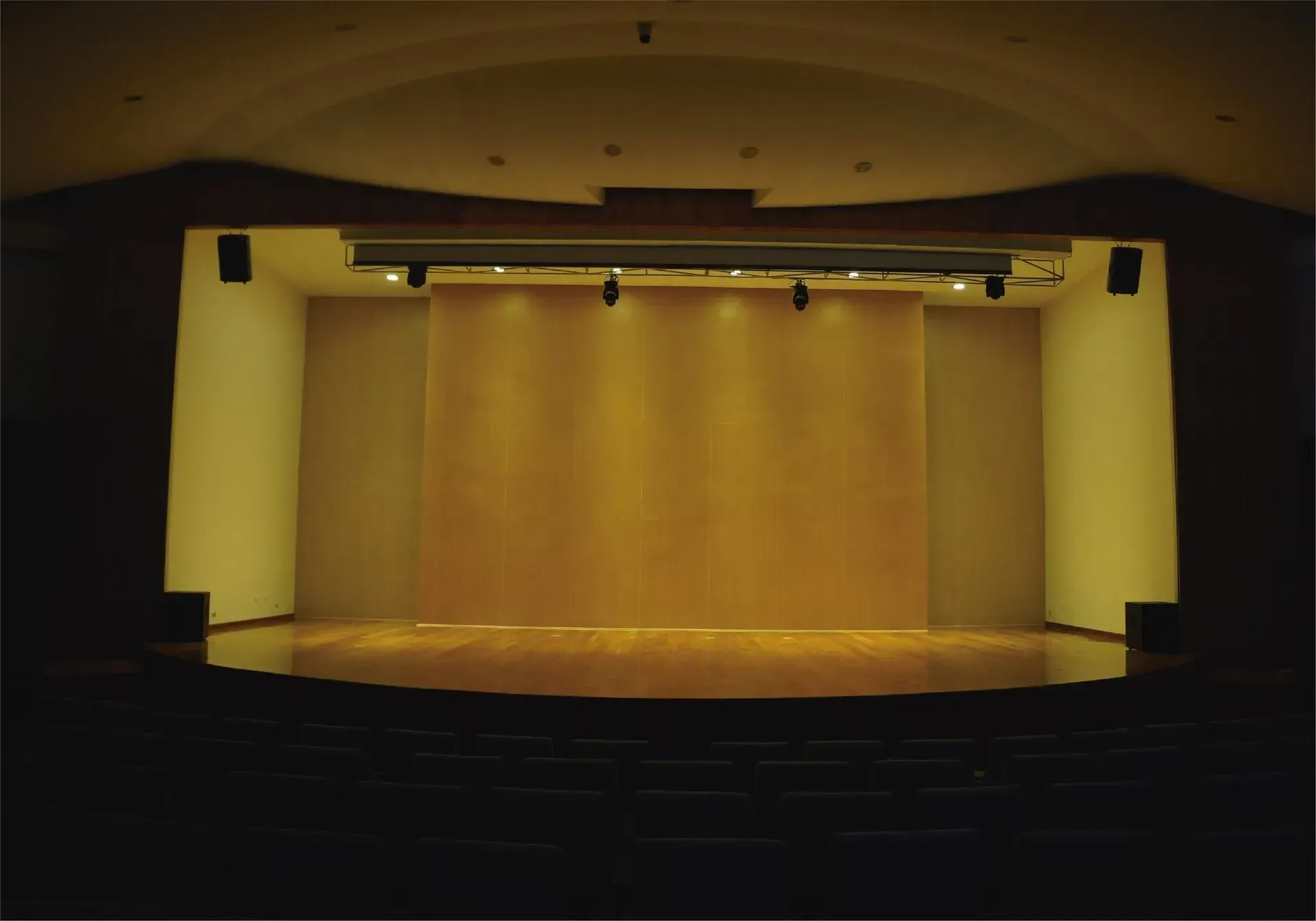 Escenario de teatro vacío, fondo de madera, iluminación cálida y asientos oscuros.