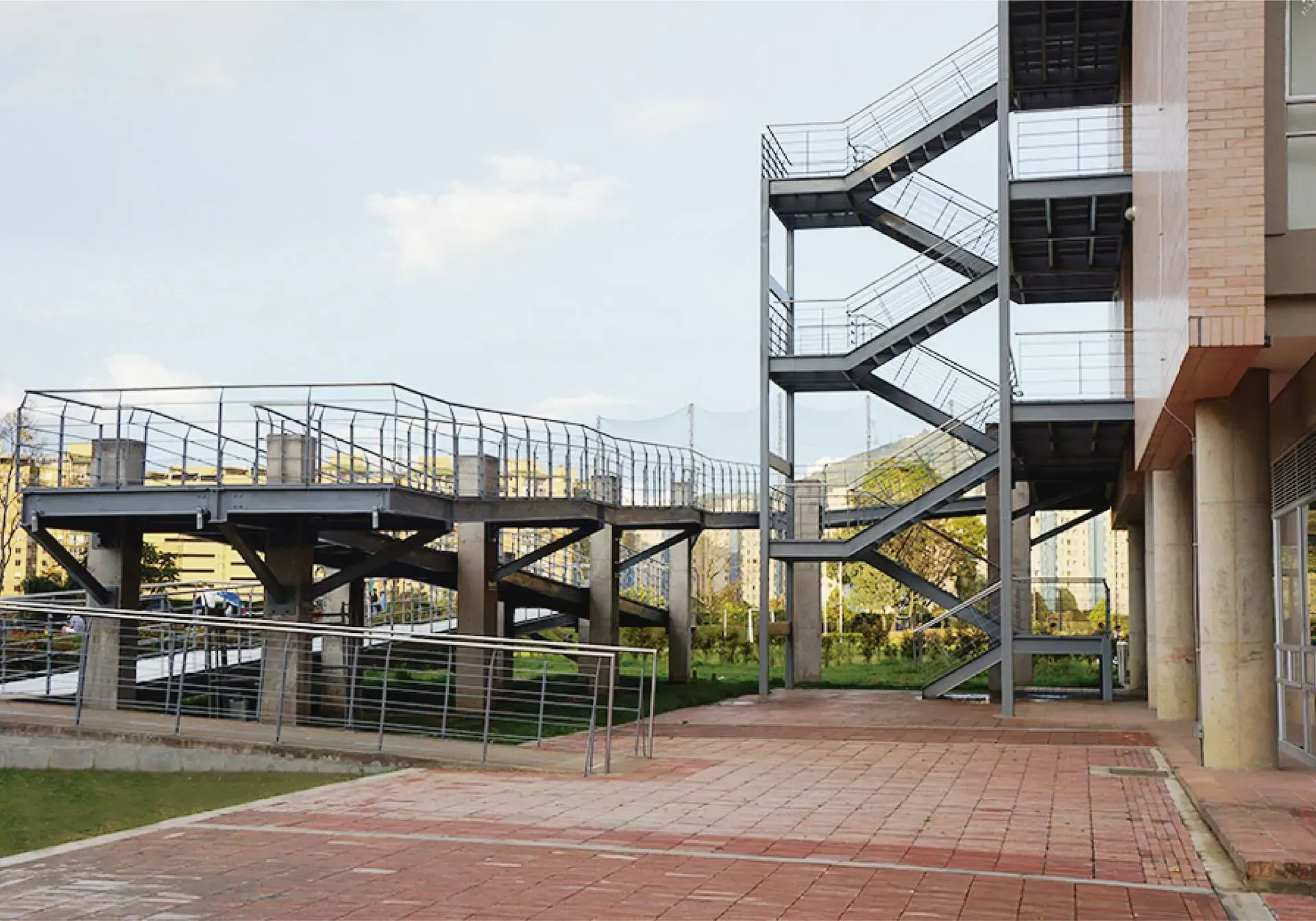 Vista exterior de una escalera metálica y una pasarela elevada junto a un edificio. Patio de ladrillo en primer plano.