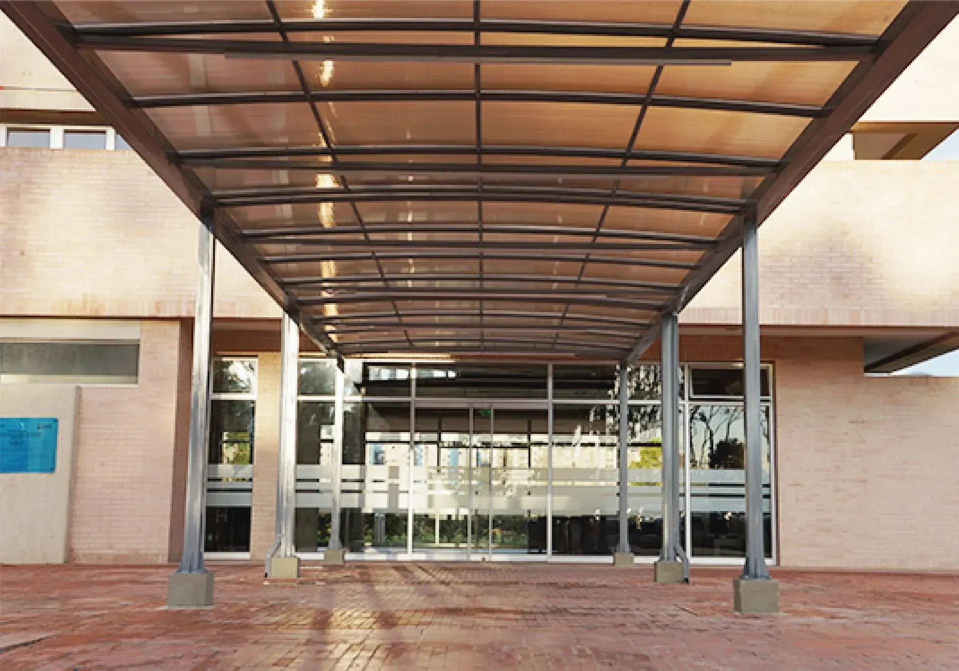 Pasarela cubierta que conduce a la entrada de un edificio con puertas de cristal. Techo y soportes marrones, suelo de ladrillo rojo.