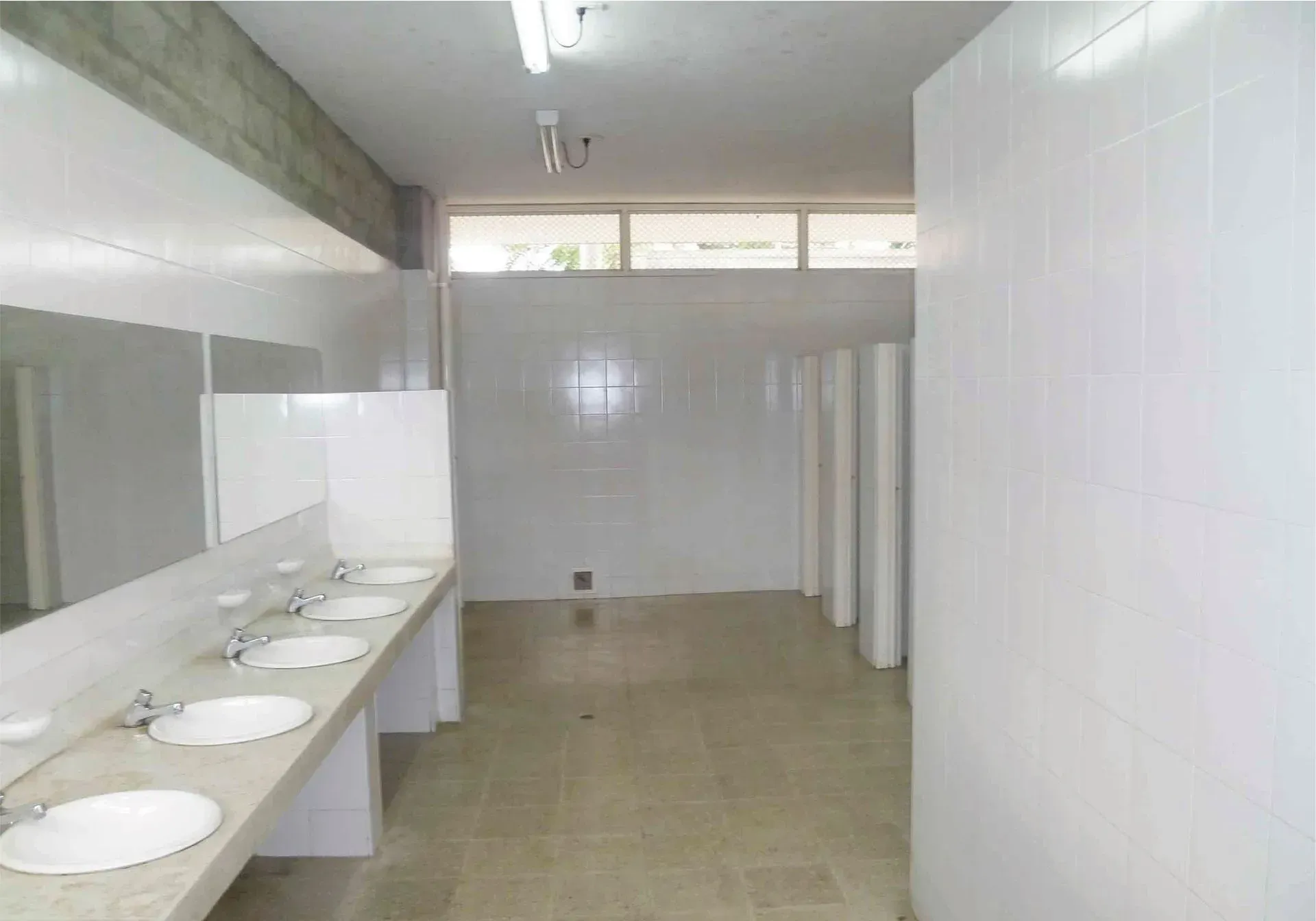 Baño público con paredes de azulejos blancos y lavabos, cubículos a la derecha.