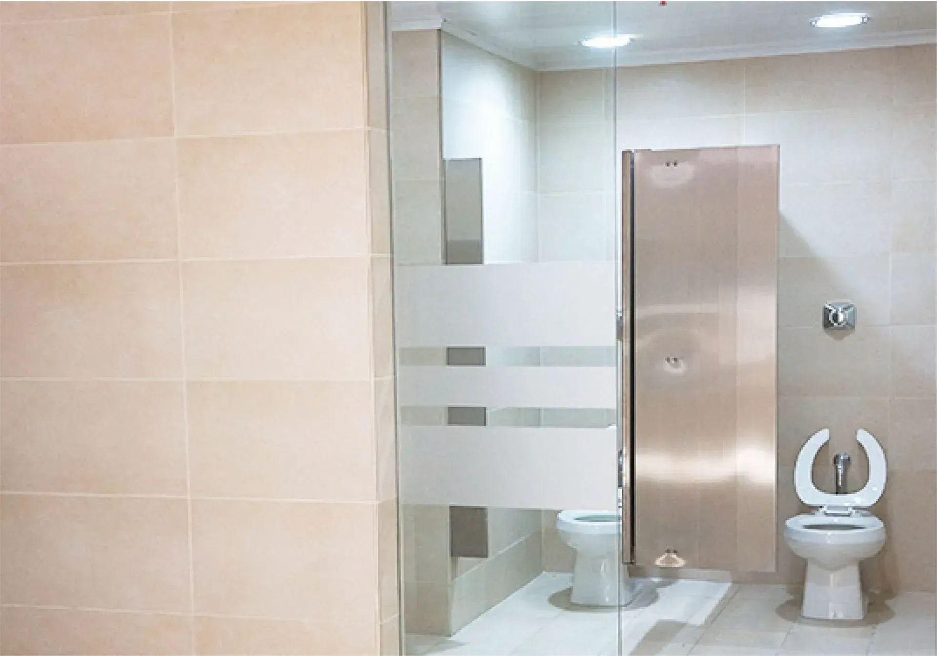 Baño público con dos sanitarios y cabina de acero inoxidable, paredes de azulejo beige, puerta de vidrio.