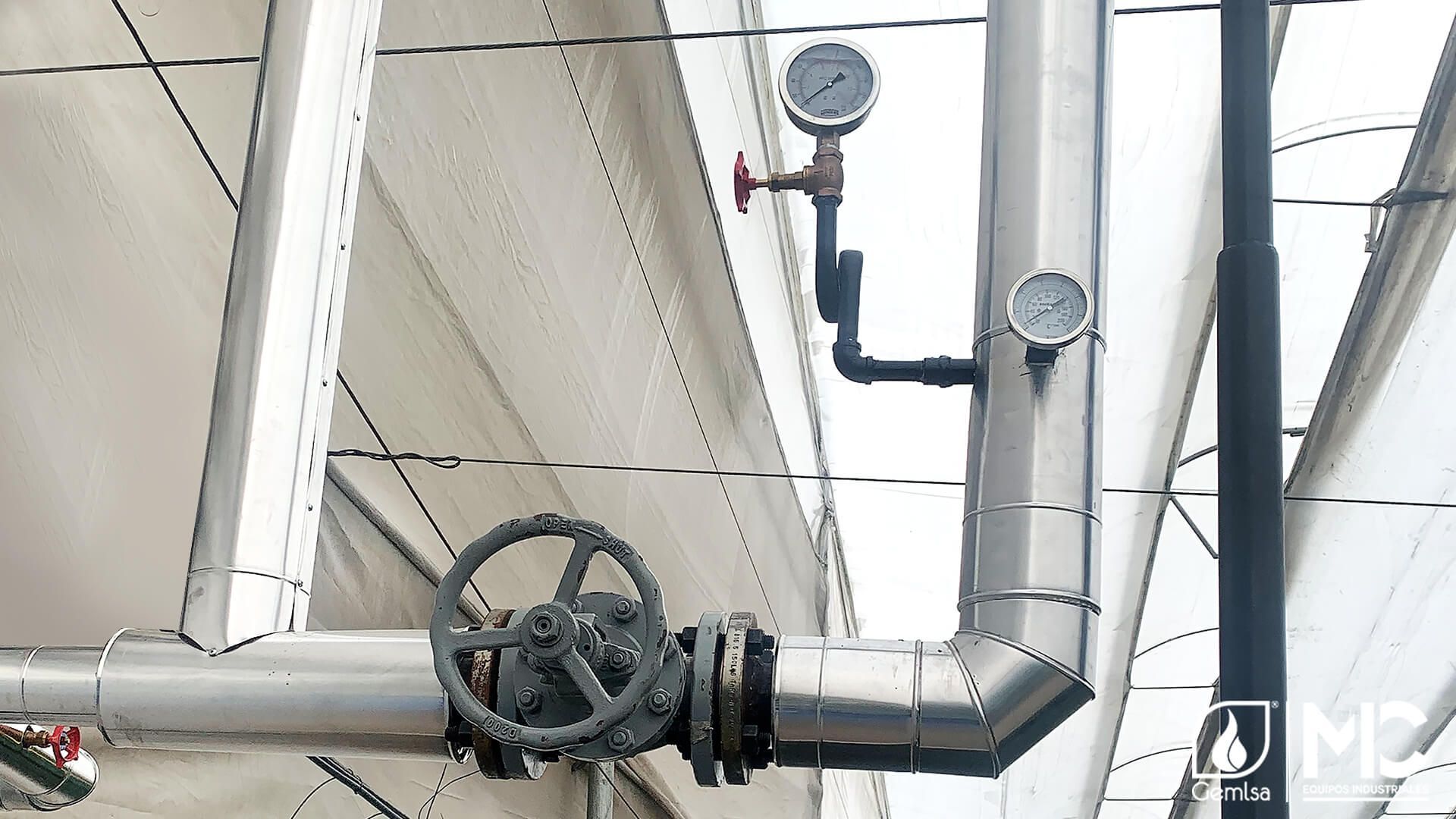 Tubos y medidores plateados con válvula, probablemente para uso industrial, sobre una estructura de edificio blanca.