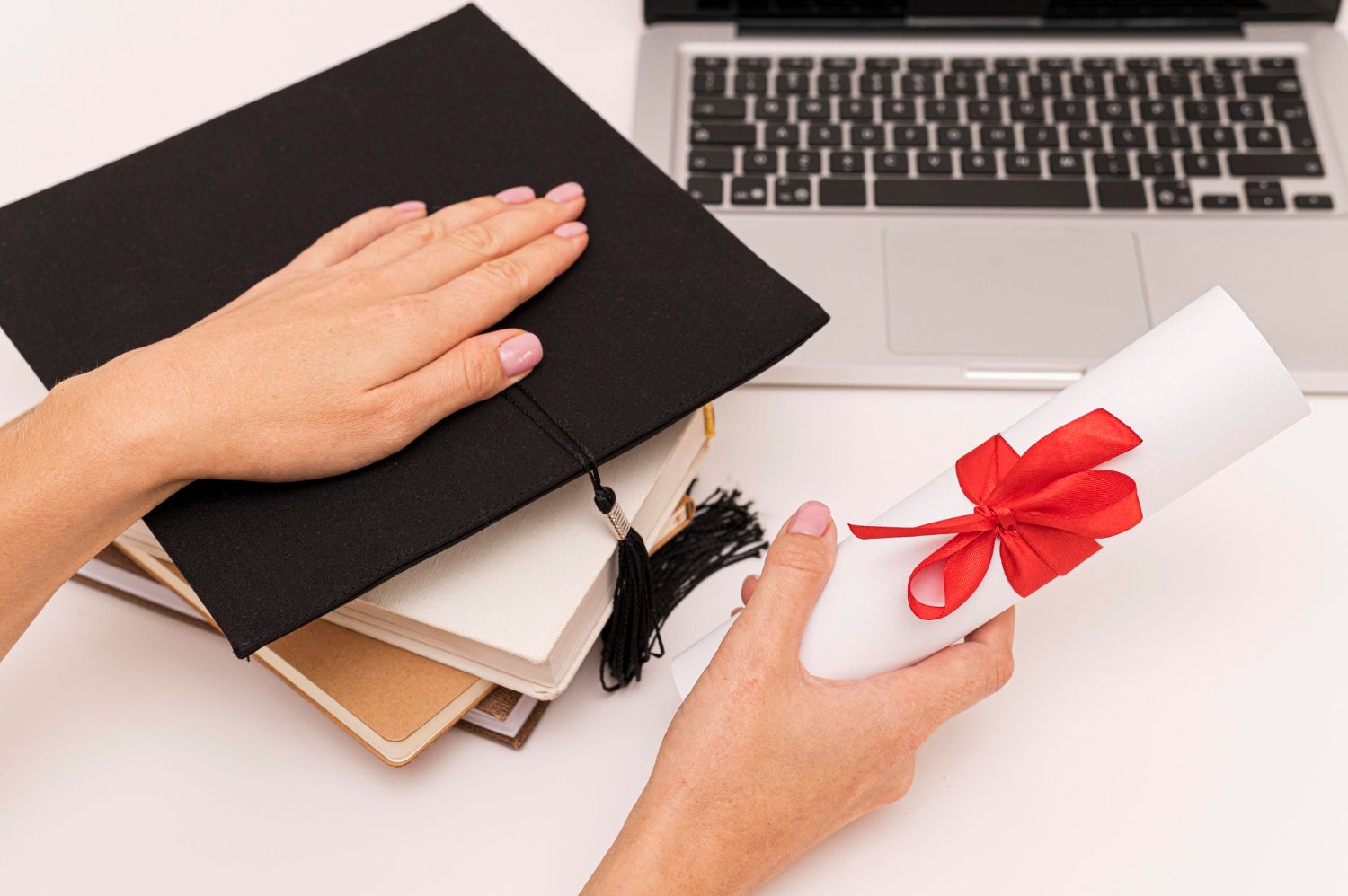 Persona sosteniendo un diploma atado con una cinta roja junto a una gorra de graduación y una computadora portátil .