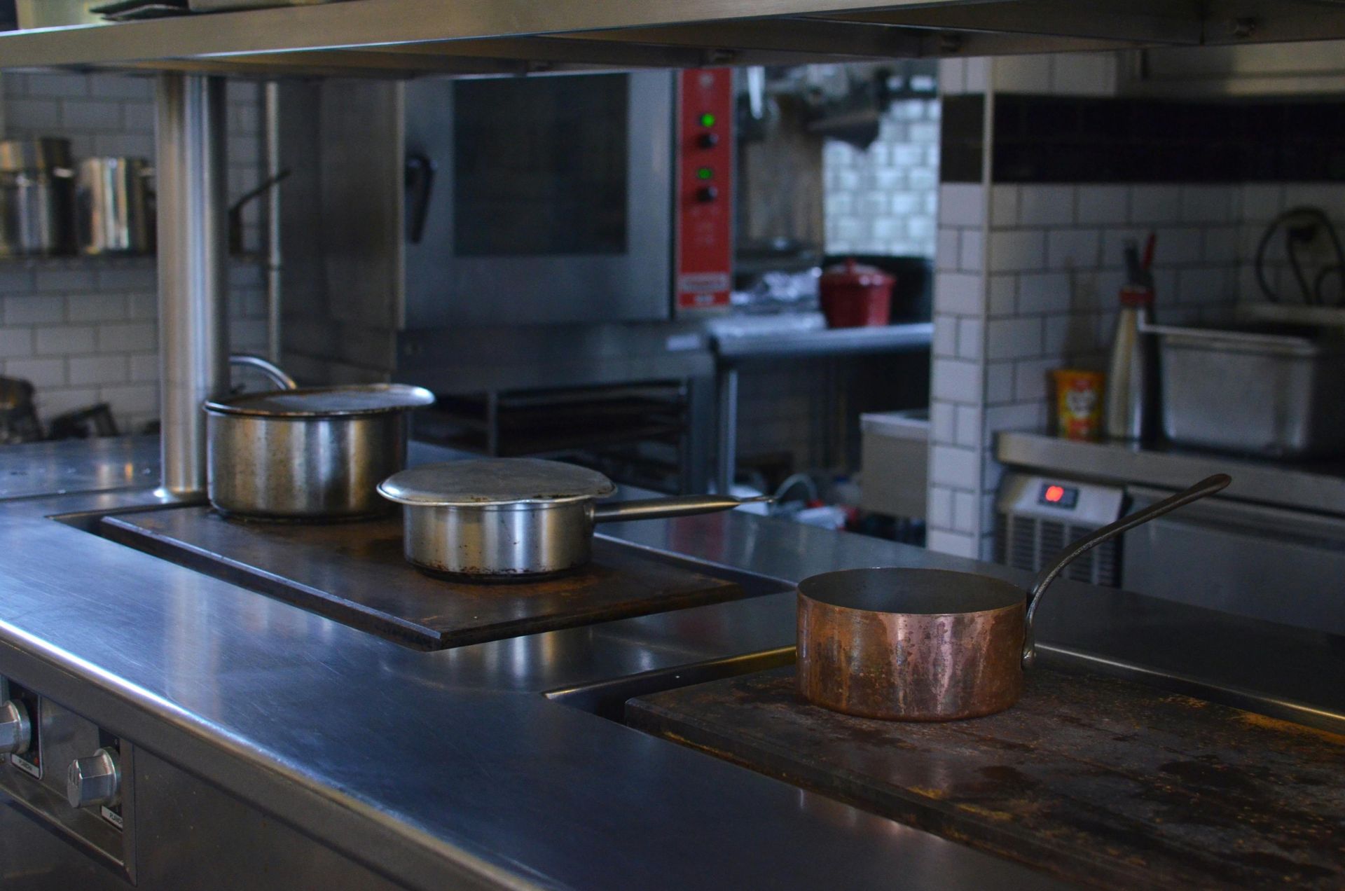Cocina comercial con tres ollas sobre hornilla, de acero inoxidable y cobre, con equipamiento al fondo.
