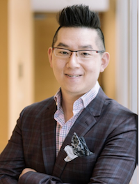 A man in a suit and glasses is leaning against a wall with his arms crossed.