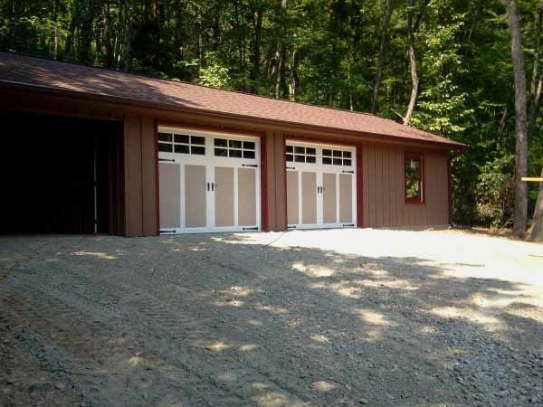 A garage with two garage doors is surrounded by trees and gravel.