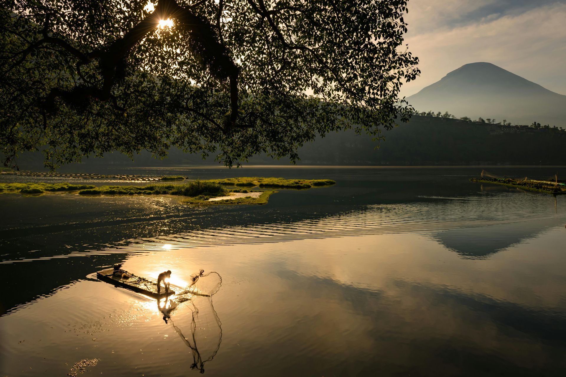 Sunlight reflecting on a calm lake with a mountain silhouette in the background, framed by tree branches.