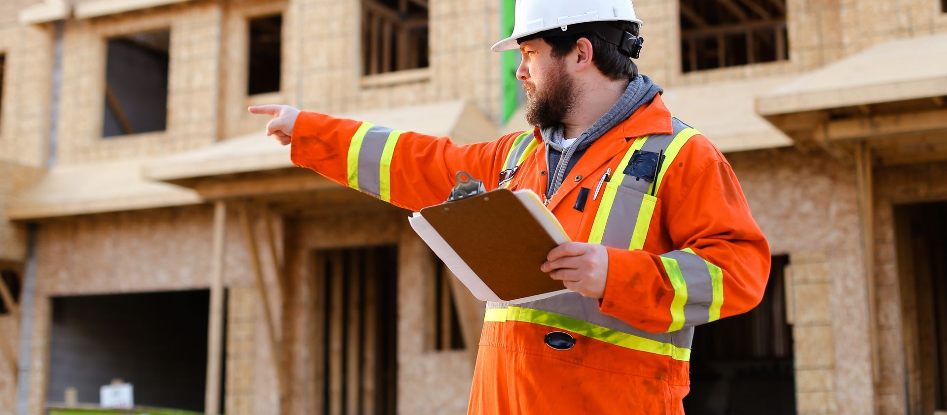 Construction worker in orange safety suit points toward unfinished buildings while holding a clipboard.