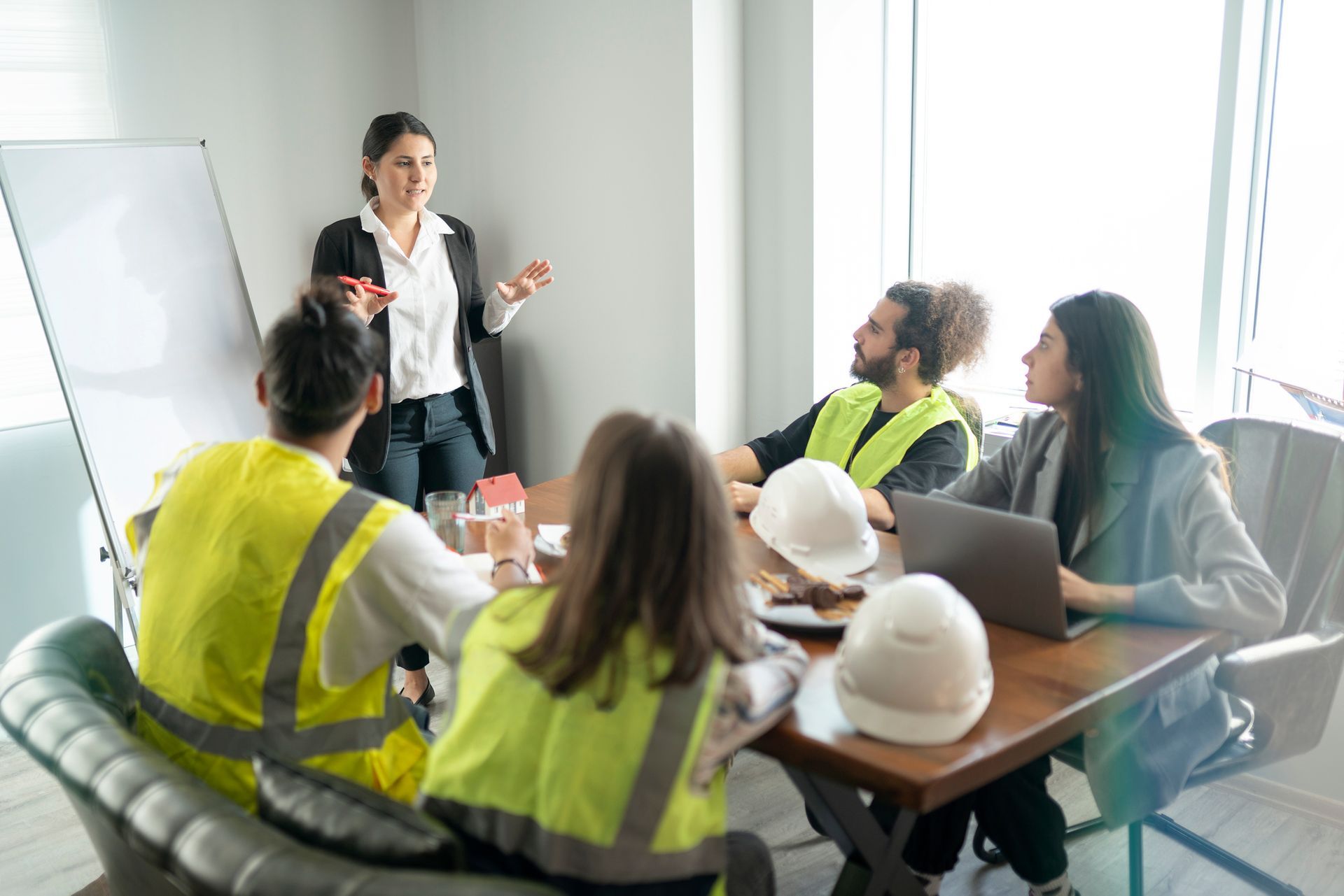 Woman presenting to a team at a conference table, some wearing safety vests and hard hats, in a well-lit office.