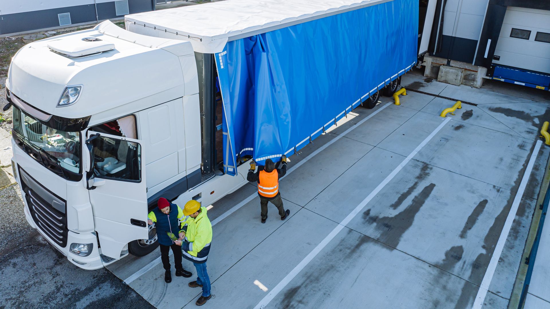 Truck being loaded at a warehouse dock, workers in safety vests adjusting blue tarp, exterior.
