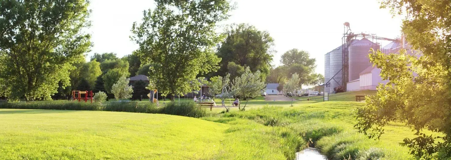 A sunny grassy park with trees, a small stream, and large metal grain silos in the distance.