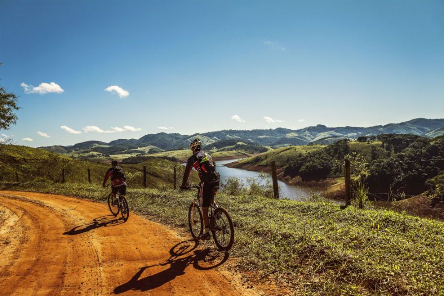 Two cyclists ride mountain bikes along a scenic dirt path overlooking a lake and rolling hills under a clear blue sky.