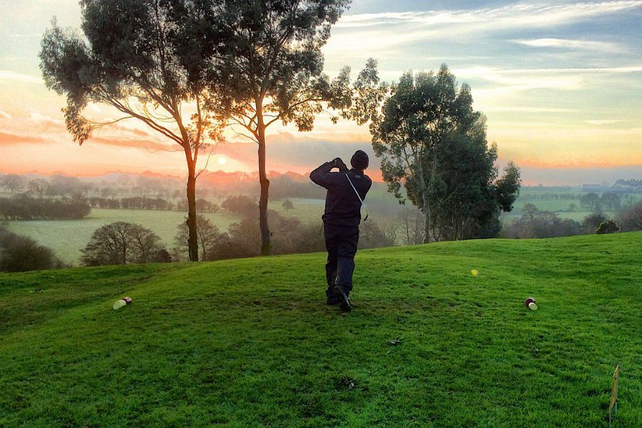 A person swings a golf club on a grassy hill during a misty, golden sunrise.
