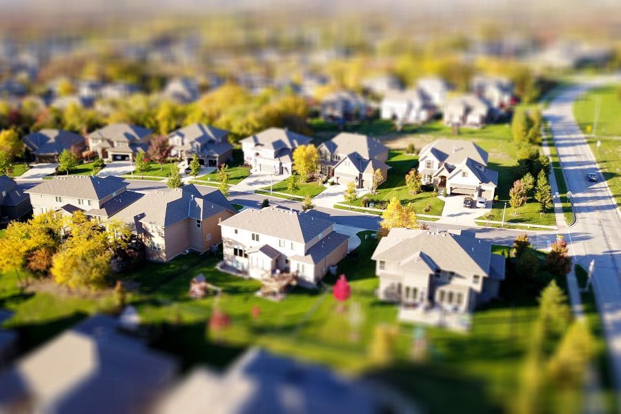 An aerial view of a suburban neighborhood with houses, green lawns, and trees, captured with a tilt-shift focus effect.