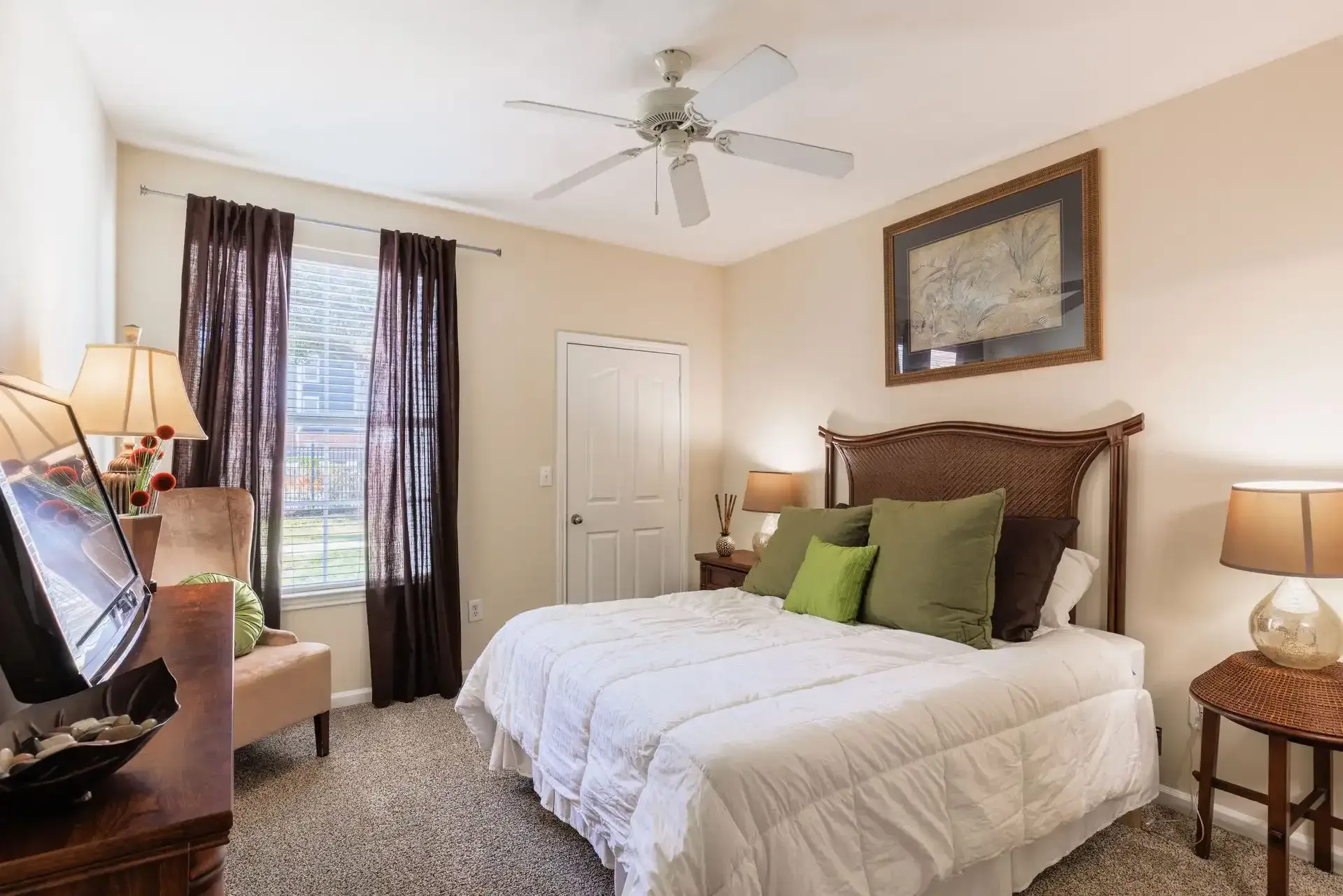 Bedroom with bed, chair, window, and artwork; neutral color scheme, dark brown curtains.