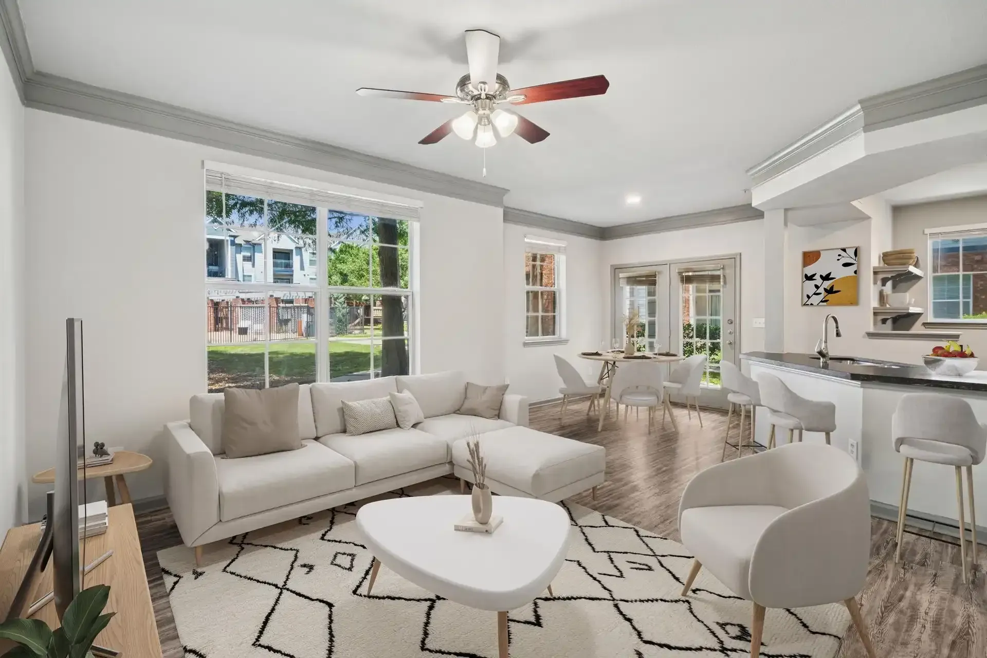 Spacious living room with a white sectional, coffee table, and rug. Large windows overlook a green space.