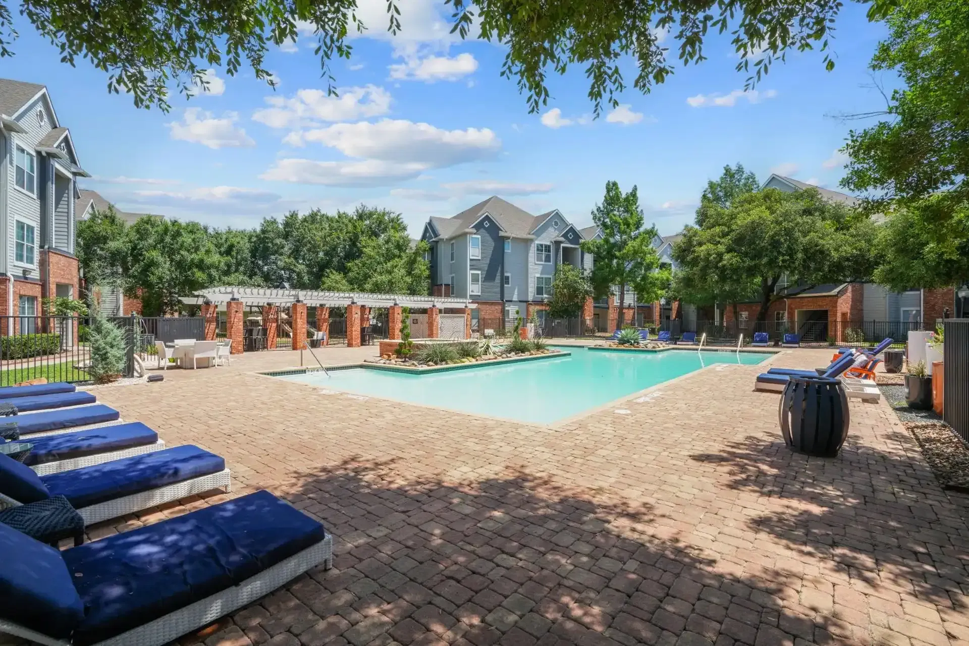 Pool area with lounge chairs, blue water, and apartment buildings under a blue sky.