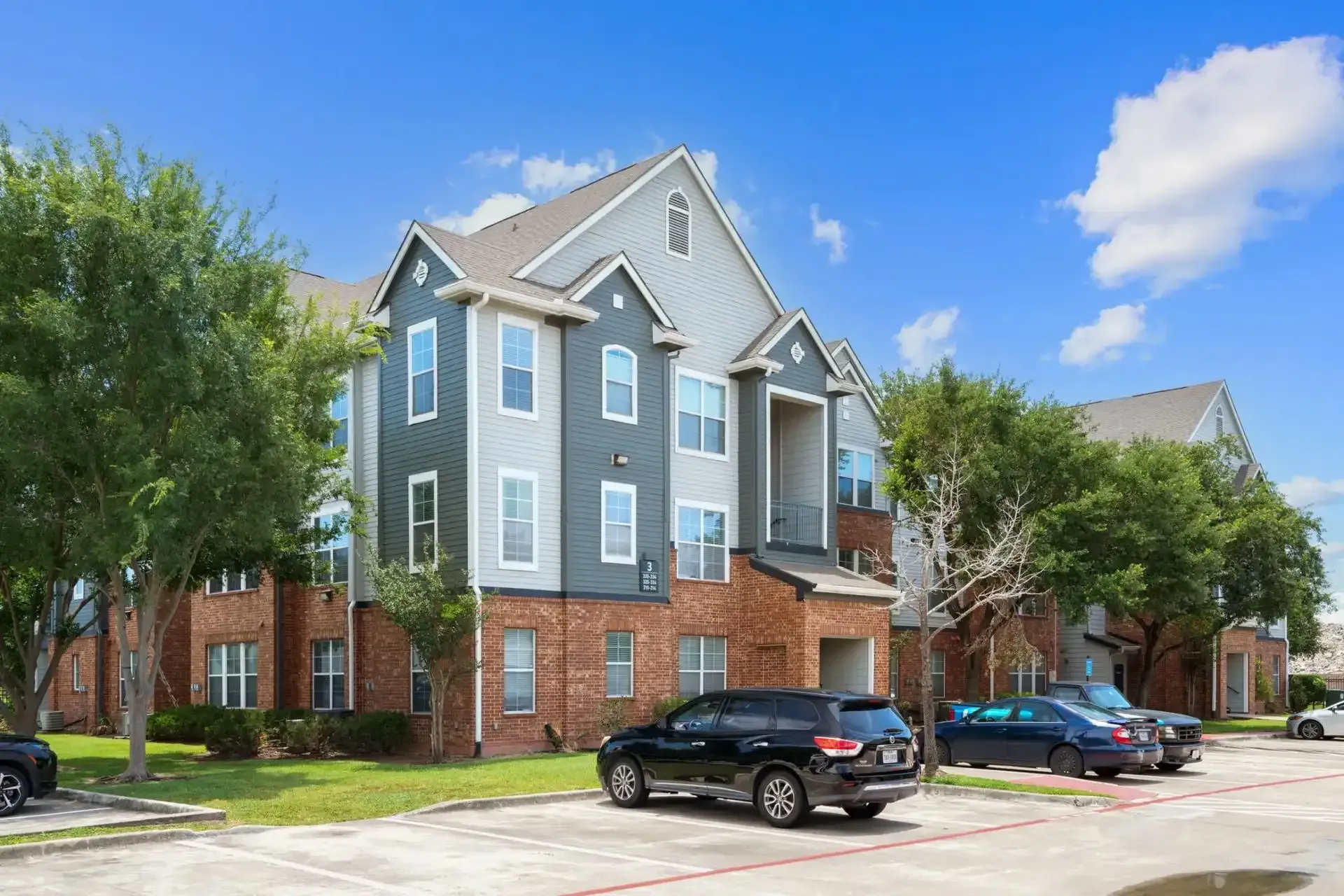 Apartment building exterior with blue siding and brick, parked cars on a sunny day.