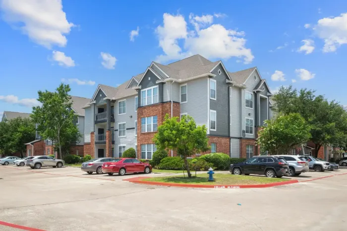 Apartment complex exterior, gray siding, brick accents, parked cars in lot, blue sky.