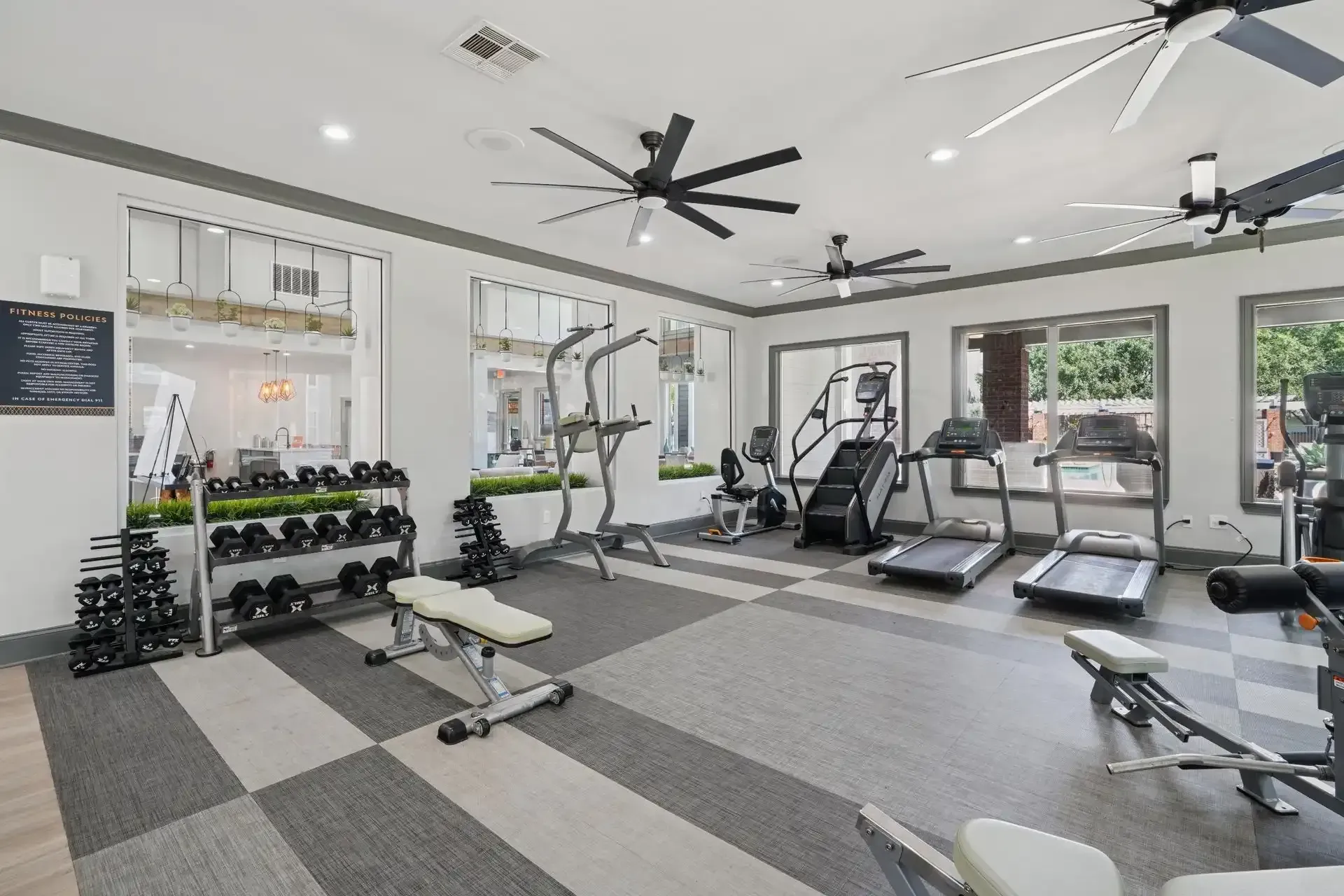 Gym with weights, machines, and fans, featuring a checkerboard floor, windows, and neutral colors.
