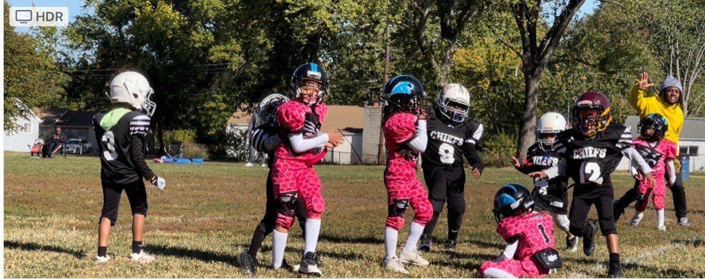 Youth football players in black, pink, and white uniforms on a field with trees.