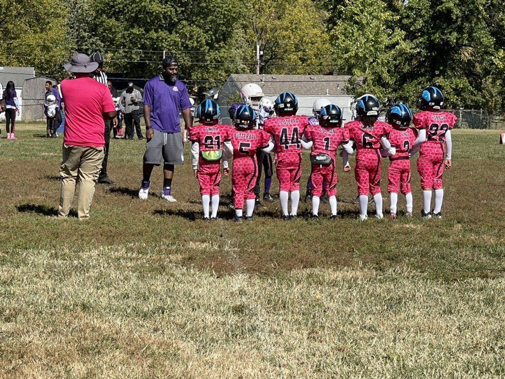 Youth football team huddles on a grassy field, wearing red and black uniforms, with two coaches.