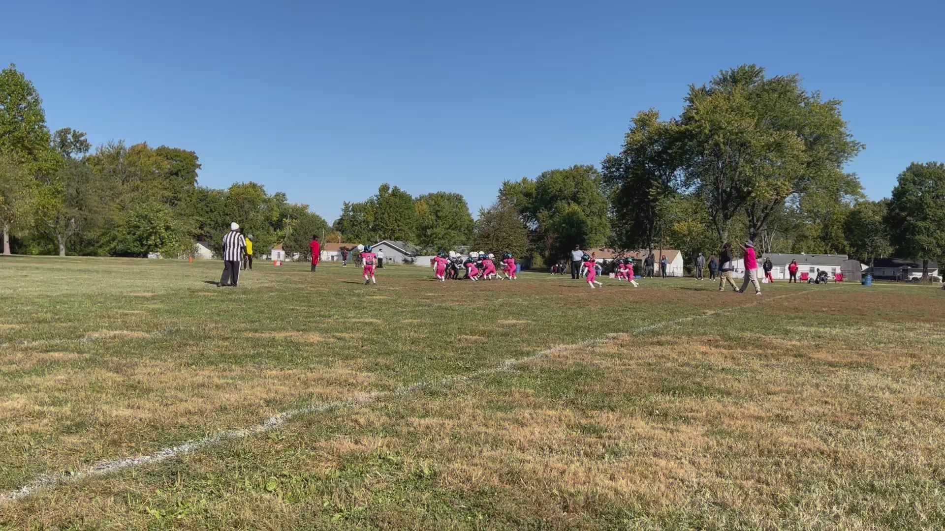 Children in pink uniforms play football on a grassy field under a blue sky.