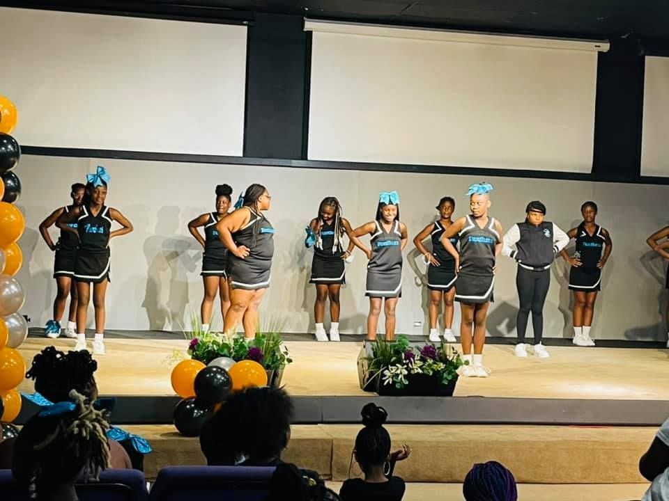 Cheerleaders on stage in black and teal uniforms, performing. Balloons and flowers decorate the stage. Audience in the foreground.
