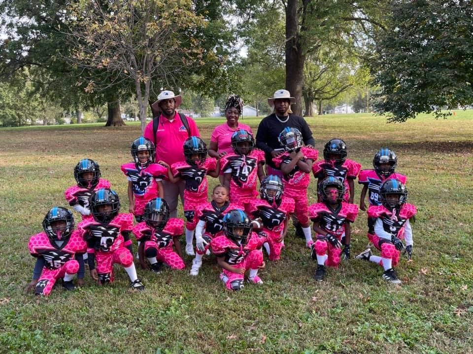 Youth football team in pink and black uniforms poses on a grassy field with coaches.