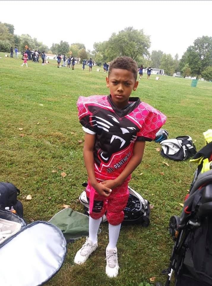 Young boy in pink and black football uniform stands on a grassy field.