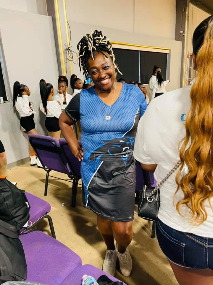Smiling woman in blue dress stands, hands on hips, near a group of dancers and another woman with long hair.