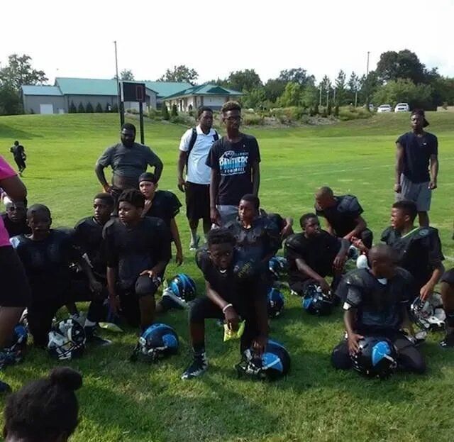 Youth football team kneels on a green field with coaches and helmets.