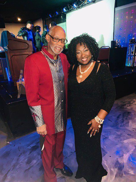 Man in red suit and woman in black dress smiling together at an event.