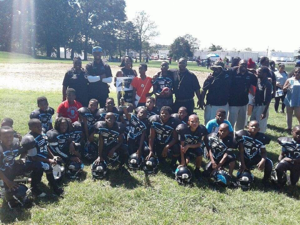 Youth football team poses on a sunny field with coaches and spectators. Players wear black and teal uniforms.
