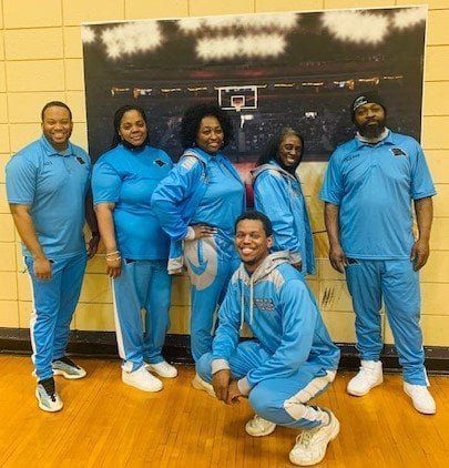 Group of seven people in blue tracksuits pose in front of a basketball court backdrop.