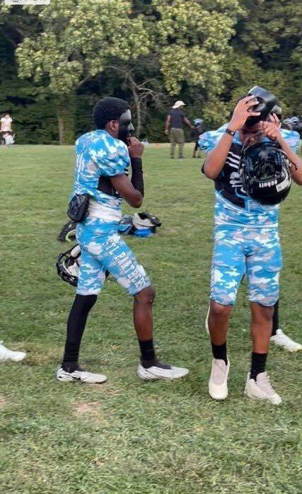 Two football players in blue camouflage uniforms on a grassy field, one wearing a helmet.
