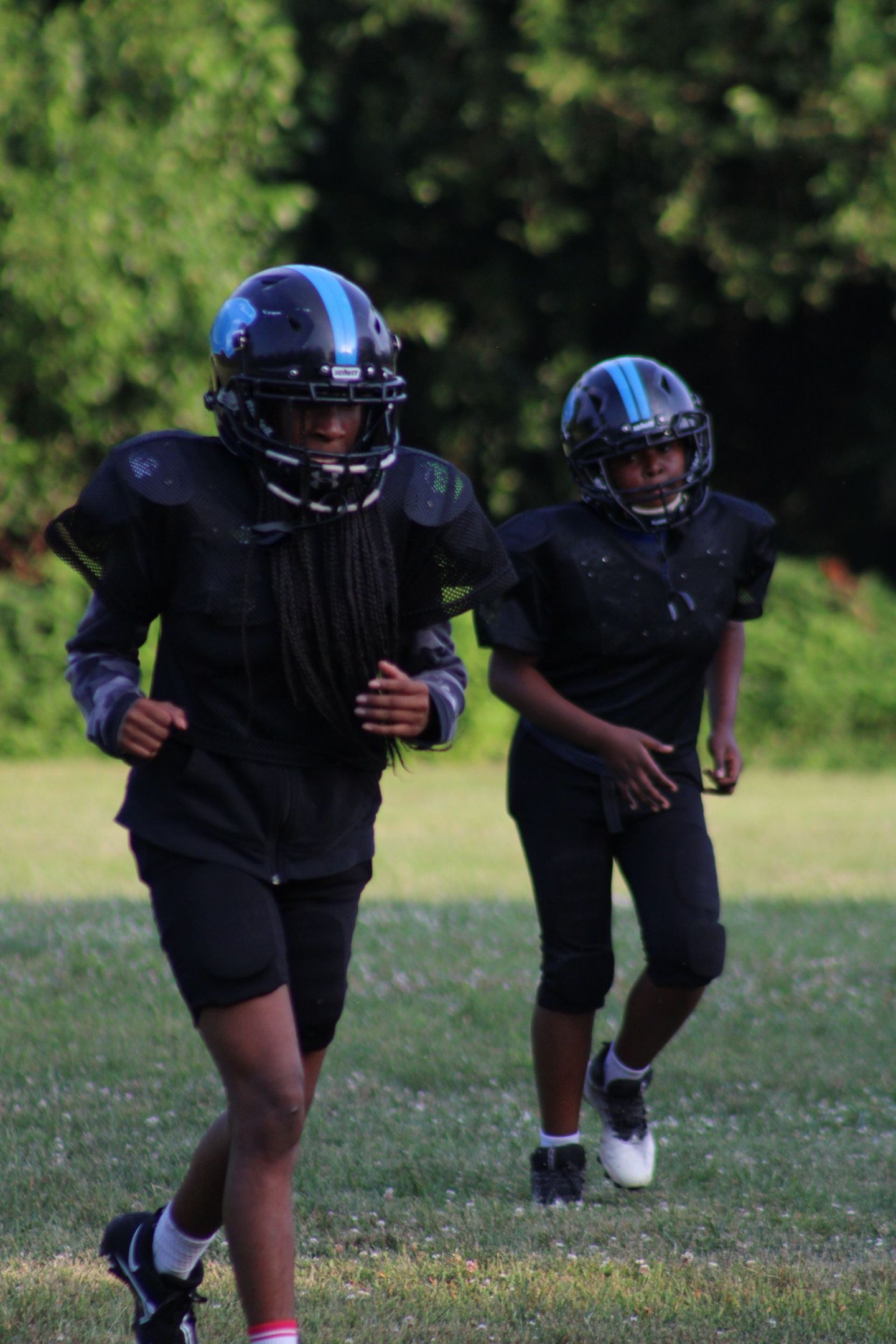Two young football players in black uniforms and helmets with blue stripes running on a grassy field.