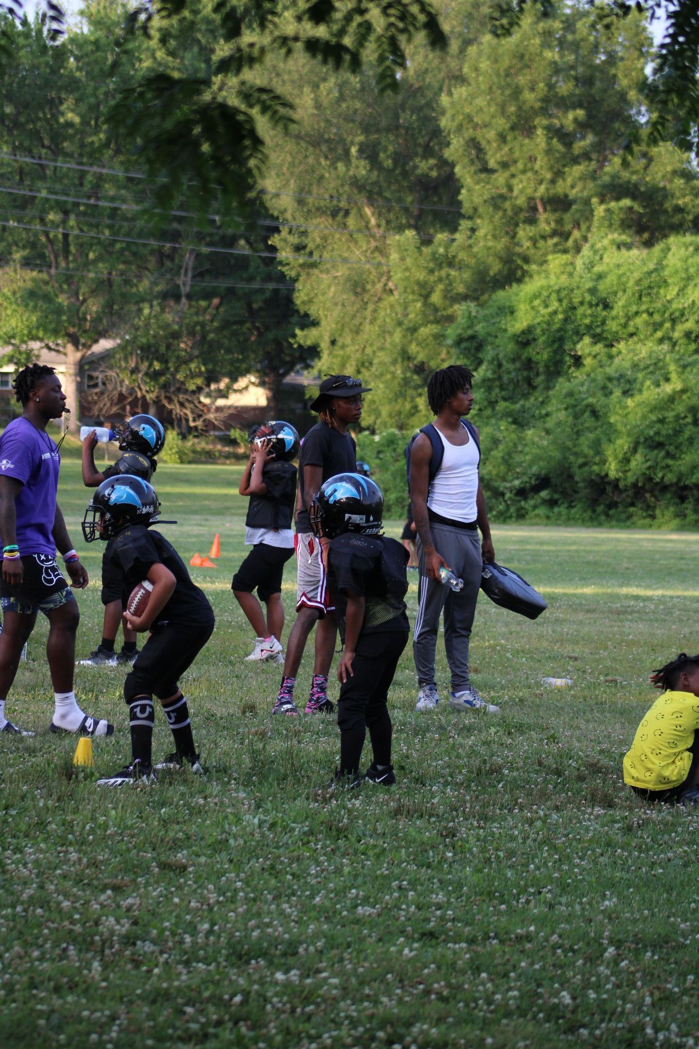 Youth football practice in a grassy park. Players in black uniforms, coaches watching, sunny day.