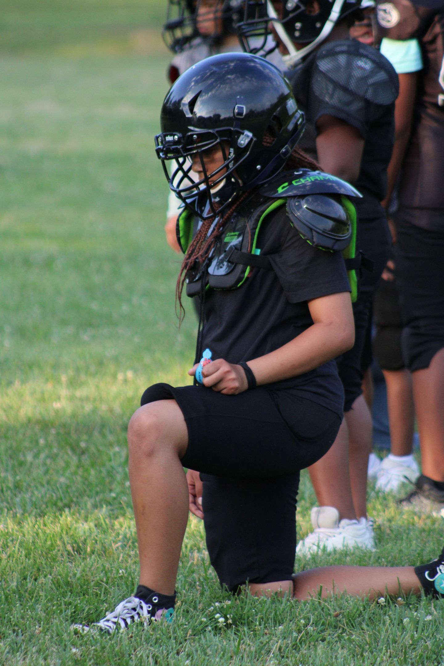 A football player in black uniform, kneeling on a grassy field, wearing a black helmet and shoulder pads.