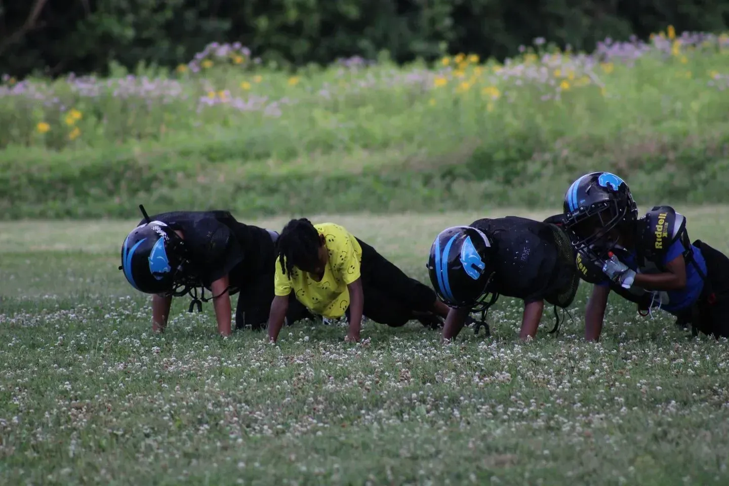Football players doing push-ups on a grassy field. One player in a yellow shirt, others in blue helmets.