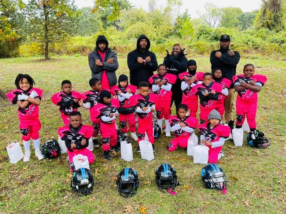 Youth football team in pink and black uniforms, posing with coaches outdoors, arms crossed.